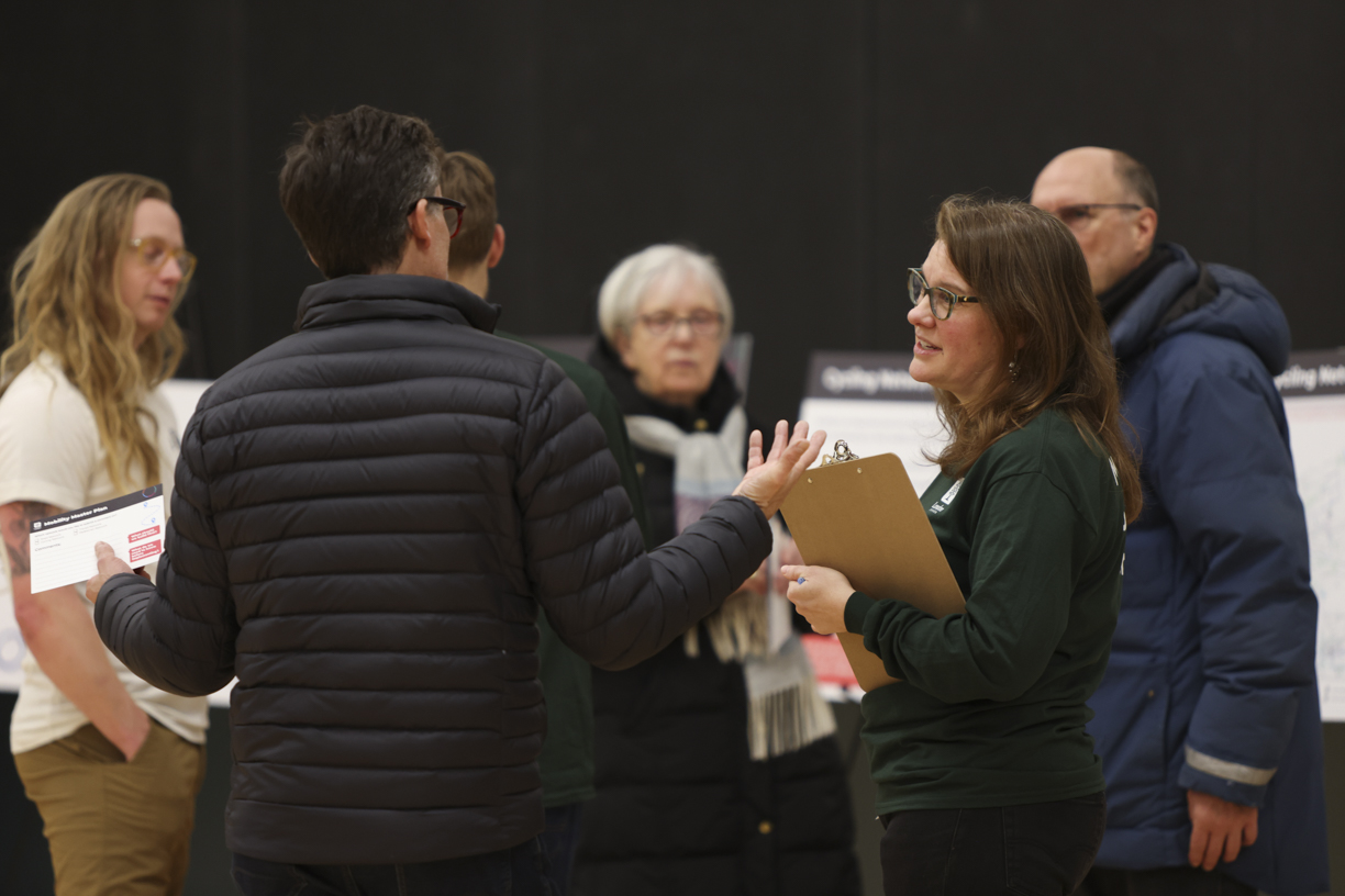 A woman in green with a clipboard is talking with a man in a jacket with his arms open and people looking on in the background at a public meeting.